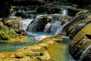 Tropical jungle waterfall in Thailand surrounded by lush green rainforest, moss-covered rocks, dense vegetation, and flowing water. The natural cascade creates a peaceful and refreshing scene © Yuliia