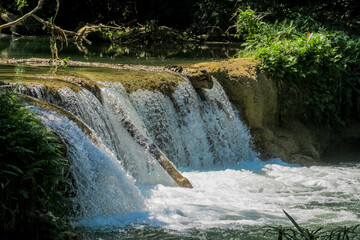 Tropical jungle waterfall in Thailand surrounded by lush green rainforest, moss-covered rocks, dense vegetation, and flowing water. The natural cascade creates a peaceful and refreshing scene © Yuliia