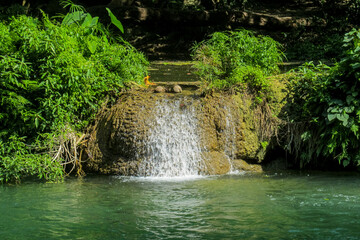 Tropical jungle waterfall in Thailand surrounded by lush green rainforest, moss-covered rocks, dense vegetation, and flowing water. The natural cascade creates a peaceful and refreshing scene © Yuliia