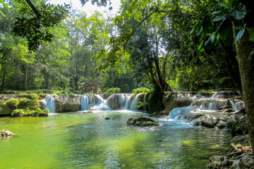 Tropical jungle waterfall in Thailand surrounded by lush green rainforest, moss-covered rocks, dense vegetation, and flowing water. The natural cascade creates a peaceful and refreshing scene © Yuliia