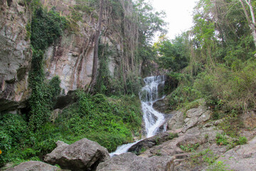 Tropical jungle waterfall in Thailand surrounded by lush green rainforest, moss-covered rocks, dense vegetation, and flowing water. The natural cascade creates a peaceful and refreshing scene © Yuliia