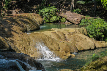 Tropical jungle waterfall in Thailand surrounded by lush green rainforest, moss-covered rocks, dense vegetation, and flowing water. The natural cascade creates a peaceful and refreshing scene © Yuliia