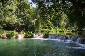 Tropical jungle waterfall in Thailand surrounded by lush green rainforest, moss-covered rocks, dense vegetation, and flowing water. The natural cascade creates a peaceful and refreshing scene © Yuliia