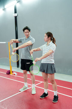 Asian male badminton coach teaching young woman proper racket grip technique on indoor court