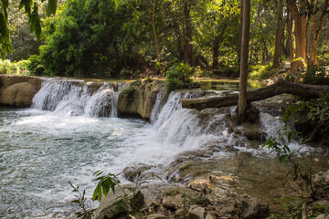 Tropical jungle waterfall in Thailand surrounded by lush green rainforest, moss-covered rocks, dense vegetation, and flowing water. The natural cascade creates a peaceful and refreshing scene © Yuliia