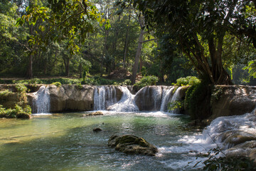 Fototapeta premium Tropical jungle waterfall in Thailand surrounded by lush green rainforest, moss-covered rocks, dense vegetation, and flowing water. The natural cascade creates a peaceful and refreshing scene