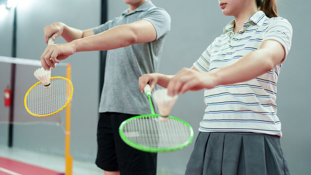 Asian coach guiding young woman holding badminton racket and shuttlecock on indoor court