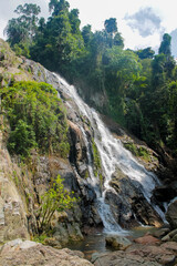 Tropical jungle waterfall in Thailand surrounded by lush green rainforest, moss-covered rocks, dense vegetation, and flowing water. The natural cascade creates a peaceful and refreshing scene © Yuliia