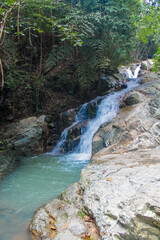 Tropical jungle waterfall in Thailand surrounded by lush green rainforest, moss-covered rocks, dense vegetation, and flowing water. The natural cascade creates a peaceful and refreshing scene © Yuliia