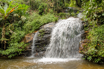 Tropical jungle waterfall in Thailand surrounded by lush green rainforest, moss-covered rocks, dense vegetation, and flowing water. The natural cascade creates a peaceful and refreshing scene © Yuliia
