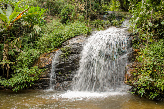 Tropical jungle waterfall in Thailand surrounded by lush green rainforest, moss-covered rocks, dense vegetation, and flowing water. The natural cascade creates a peaceful and refreshing scene