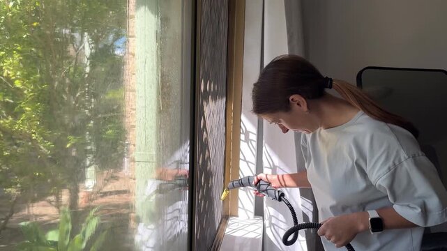 Young woman cleaning a dusty glass window pane and frame with a portable steam cleaner appliance inside a bright residential home