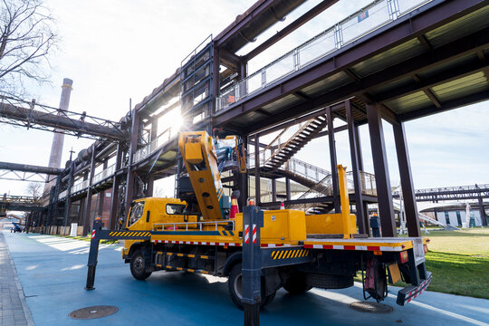 A bright yellow aerial work truck parks firmly on flat ground inside an old industrial zone.