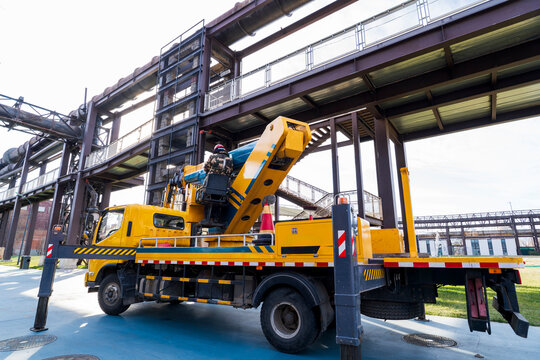 A bright yellow aerial work truck parks firmly on flat ground inside an old industrial zone.
