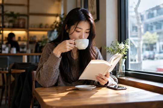 Young woman enjoying coffee while reading a book by a cafe window, representing peaceful leisure, reading, relaxation, and modern everyday lifestyle.
