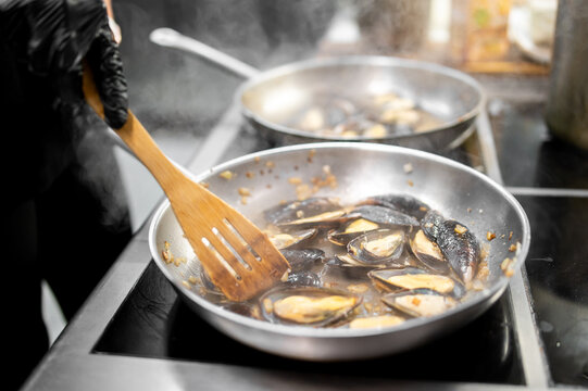 Chef cooking fresh mussels in white wine sauce in stainless steel pan on induction stove.