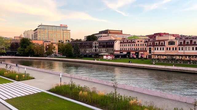 Riverbank with buildings and park on sunset in city. Svisloch River embankment. Landscaped waterfront with pedestrian and historic architecture of city center. Belarus, Minsk, September 16, 2026