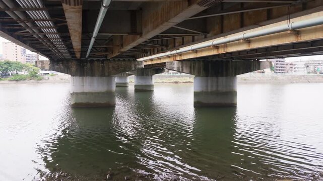 Low angle view under a rusty steel bridge over the Ota River in Hiroshima, Japan. Calm water ripples reflect concrete piers on an overcast day. Urban river background for documentary and travel.