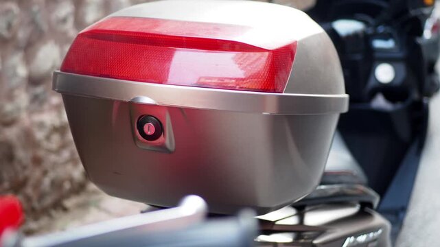 Close-up of a parked silver scooter's top case or trunk, featuring a prominent red reflector and lock mechanism