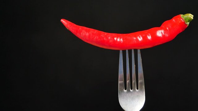 A single red hot chili pepper balancing on a metal fork, rotating slowly against a black background, capturing its vibrant color and smooth texture in a dynamic motion clip