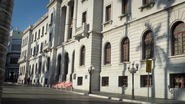 Historic white municipal building of Tampico facing main square illuminated by soft sunrise light with palm tree shadows on walls and city street morning movement