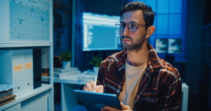 Young Caucasian male programmer use tablet for code design and plan in dark home office at night. AI technology, cybersecurity, software developer, freelance innovation, digital data network concept.