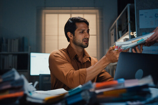 Overworked Latin businessman receiving extra paperwork from boss at desk in dark night office. Working late overload, deadline burnout, heavy overtime stress, corporate business problem concept.
