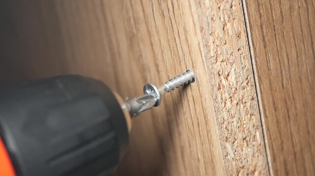 Electric screwdriver driving metal screw into wooden furniture panel for home repair and construction projects