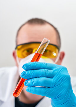 Laboratory worker holding test tube with red liquid in front, protective gloves, goggles and face mask, examining sample consistency