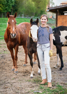 Young woman walking next to two American Paint Horses, blurred ranch with wooden boxes for animals in background