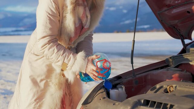 Woman pouring anti freeze windshield fluid into car on cold winter day, snow covered fields and mountains blurred background, closeup to hands holding bottle with blue liquid
