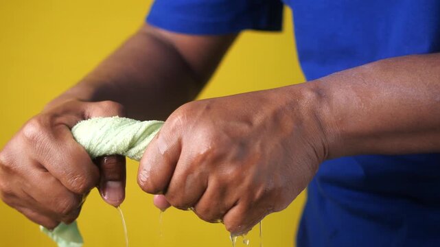 Hands squeezing water out of wet green towel against yellow background in studio shot