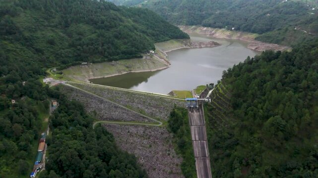 Elevated drone perspective of a massive dam structure and wide reservoir bordered by steep, forested hillsides and winding access roads