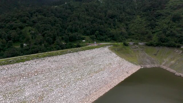 Aerial perspective of a large sloping rock-fill dam holding back a deep mountain reservoir bordered by dense green forests