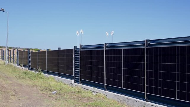 an extended vertical solar panel fence along a property line, backed by small vertical-axis wind turbines under a clear blue sky