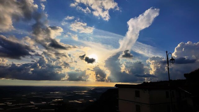 Fast moving clouds reveal radiant sun rays over a vast open plain, framed by a silhouetted house and streetlamp. Dynamic skies inspire a deep sense of natural beauty and atmospheric evolution.