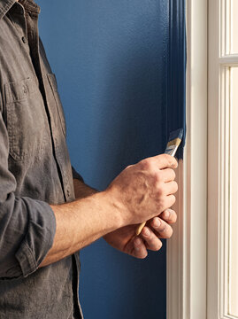 Worker painting a dark blue wall with a brush beside a window. The close-up highlights careful interior finishing, craftsmanship and professional renovation work