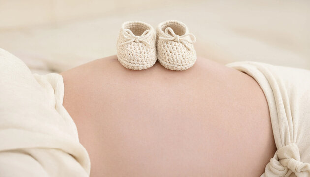 Small white baby shoes resting on the bare belly of a pregnant woman during her third trimester