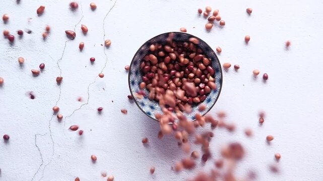 Raw peanuts falling into ceramic bowl on white surface in slow motion top view