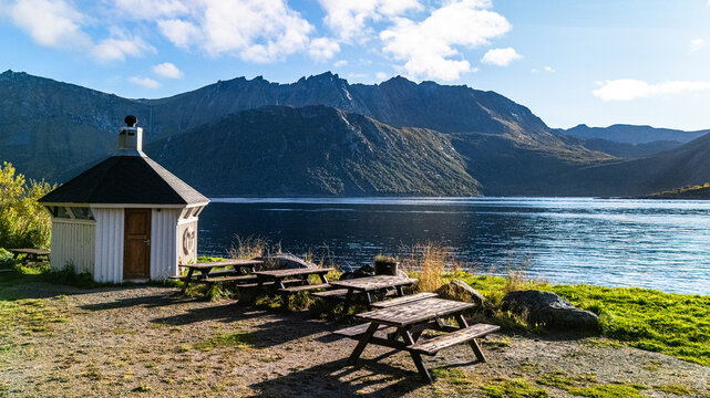 Wooden picnic tables and traditional white pavilion at a scenic fjord resting area with mountain backdrop under blue sky in Norway