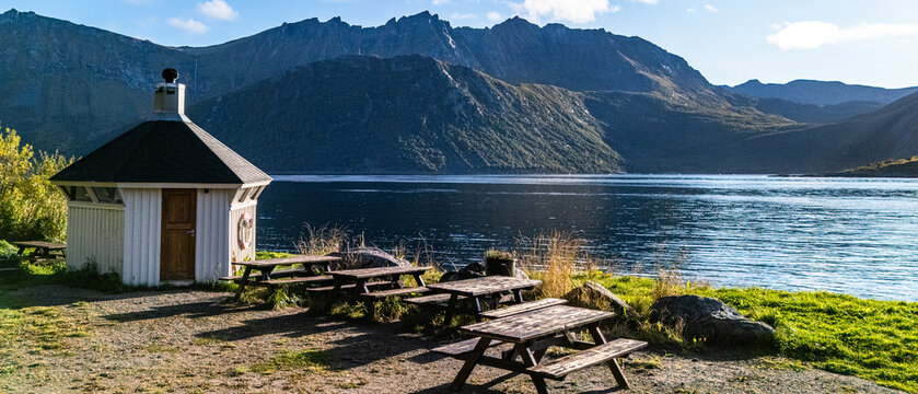 Wooden picnic tables and traditional white pavilion at a scenic fjord resting area with mountain backdrop under blue sky in Norway
