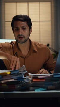 Vertical Screen : Overworked young Latin businessman working late on document large stack of paperwork files at office night. Tax audit deadline, accounting stress, corporate burnout, urgent business.
