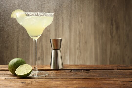 Refreshing margarita cocktail in glass, limes and jigger on wooden table, closeup. Space for text