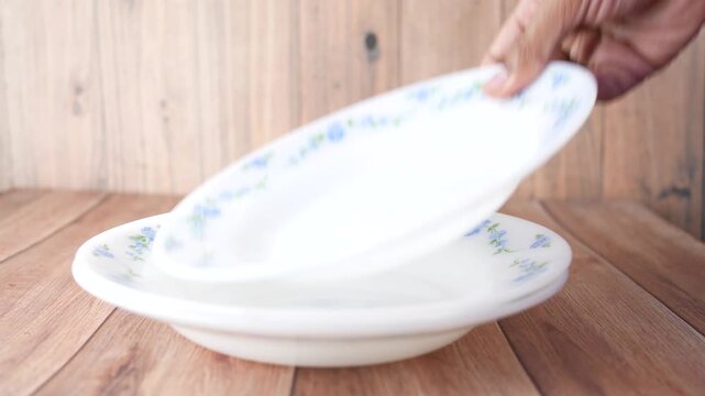 A person stacking clean, white, patterned bowls on top of each other on a wooden table, preparing for a meal or storage with gentle motion