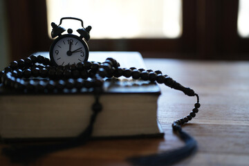 The Quran, clock, and prayer beads on a wooden table with dim lighting. Creates an atmosphere of spiritual contemplation and a sense of reverence. Reminds us of the time to pray. Muslim, Ramadan, Isla © PANCASONA CREATION