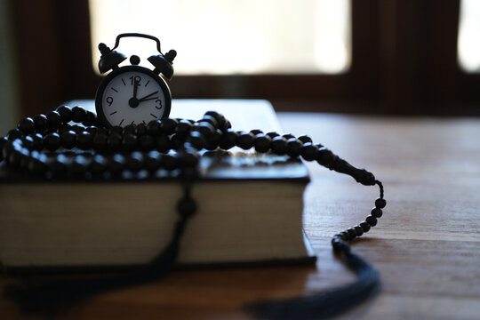 The Quran, clock, and prayer beads on a wooden table with dim lighting. Creates an atmosphere of spiritual contemplation and a sense of reverence. Reminds us of the time to pray. Muslim, Ramadan, Isla