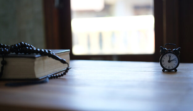 A Quran, a clock, and prayer beads on a wooden table evoke a moment of spiritual reflection and reverence. A background image suitable for Muslims, Eid al-Fitr, Eid al-Adha, and the Muharram New Year.