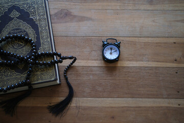 The Quran, clock, and prayer beads on a wooden table with dim lighting. Creates an atmosphere of spiritual contemplation and a sense of reverence. Reminds us of the time to pray. Muslim, Ramadan, Isla © PANCASONA CREATION