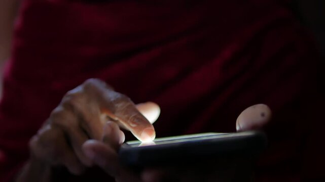 Close up of an elderly person's hands using a smartphone in a dimly lit room, focusing on the glowing screen and finger movements