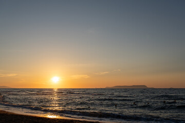Fototapeta premium Sun sets over dark rippling sea water near shoreline. Coastal view with golden sunlight reflection on waves, clear evening sky, and distant island silhouette.